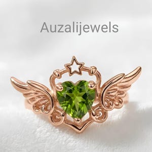 May include: Rose gold ring featuring a heart-shaped green gemstone, flanked by stylized wings. A star sits atop the heart, with the text "Auzalijewels" in the background. The ring's design is intricate and detailed.