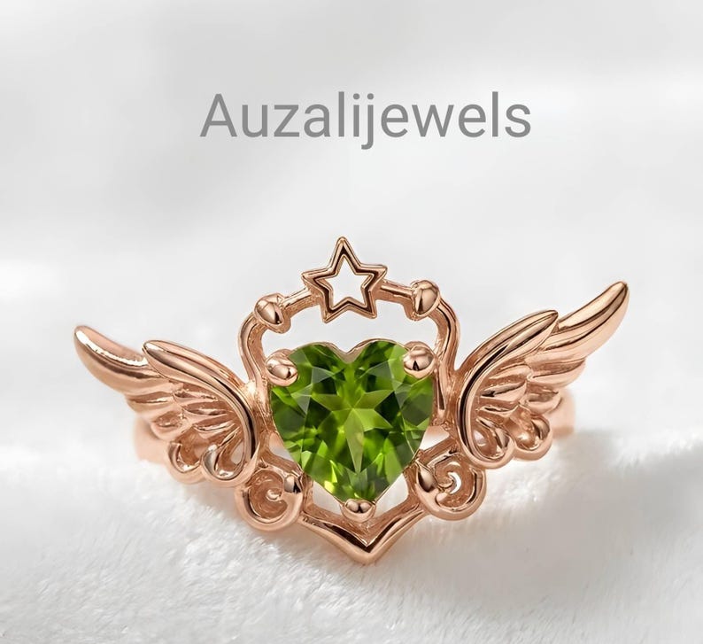 May include: Rose gold ring featuring a heart-shaped green gemstone, flanked by stylized wings. A star sits atop the heart, with the text "Auzalijewels" in the background. The ring's design is intricate and detailed.