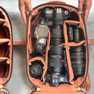 Może przedstawiać: Dwie otwarte torby na aparat. Jedna jest pusta z regulowanymi pomarańczowymi przegrodami. Druga jest wypełniona sprzętem fotograficznym, w tym korpusem aparatu, obiektywami i lampą błyskową. Torby są brązowe z pomarańczowymi wnętrzami.