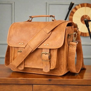 May include: A brown leather satchel with a top handle, shoulder strap, and two front pockets secured by buckles. The bag has a flap closure and metal hardware. The leather has a distressed appearance.