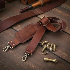 Könnte beinhalten: Ein brauner Leder-Schulterriemen mit Metallbeschlägen, darunter eine Schnalle und Verschlüsse. Mehrere Messingschrauben und ein dekoratives Metallteil liegen in der Nähe auf einer Holzoberfläche. Der Riemen hat eine gepolsterte Schulterauflage.