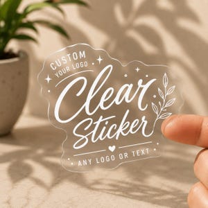 Puede incluir: Una pegatina transparente troquelada con texto impreso en blanco que dice CUSTOM YOUR LOGO, Clear Sticker y ANY LOGO OR TEXT. El diseño incluye elementos decorativos como pequeñas estrellas de cuatro puntas, un corazón, puntos y una rama.