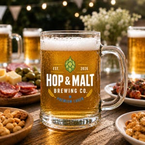 Puede incluir: Una jarra de cerveza de cristal transparente llena de cerveza rubia dorada y espuma blanca. El frente presenta un icono de l&uacute;pulo verde, el texto "EST. 2026", "HOP & MALT BREWING CO." y "PREMIUM LAGER" en blanco y azul, con un asa resistente.