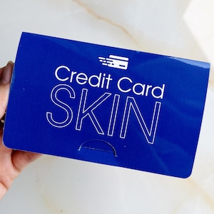 May include: A blue rectangular product with the words "Credit Card SKIN" in white. The product has a credit card icon in the upper right corner. The product is held by a person.