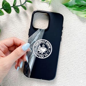 May include: Black phone case with a white sticker that reads "THE DAILY BREW COFFEE CO." featuring a coffee cup and leaves. A hand is peeling off the protective film. The case has a camera cutout and side buttons.