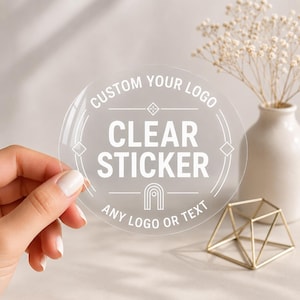 May include: A circular transparent sticker featuring white printed text that reads CUSTOM YOUR LOGO, CLEAR STICKER, and ANY LOGO OR TEXT, accompanied by geometric line art including a central arch icon and diamond shapes along the border.