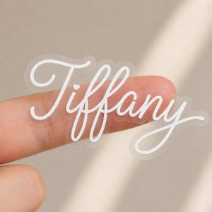 Puede incluir: Una pegatina transparente con el nombre Tiffany escrito en una fuente cursiva blanca. Las letras incluyen un pequeño icono de corazón sobre la letra i, y la pegatina tiene un borde transparente que sigue la forma del texto.