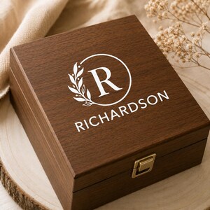 Puede incluir: Una caja cuadrada de madera marr&oacute;n oscuro con tapa abatible y un cierre de metal dorado. La parte superior de la tapa presenta un dise&ntilde;o impreso en blanco que consiste en un marco circular, una rama con hojas, la letra R y el nombre RICHARDSON.