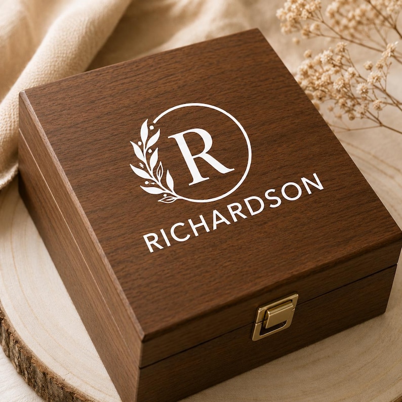 Puede incluir: Una caja cuadrada de madera marr&oacute;n oscuro con tapa abatible y un cierre de metal dorado. La parte superior de la tapa presenta un dise&ntilde;o impreso en blanco que consiste en un marco circular, una rama con hojas, la letra R y el nombre RICHARDSON.