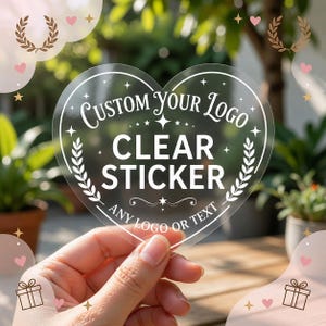 Puede incluir: Una pegatina transparente en forma de corazón con texto impreso en blanco que dice Custom Your Logo, Clear Sticker y Any Logo Or Text. El diseño incluye ramas de laurel decorativas, pequeñas estrellas y adornos alrededor del texto.