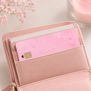 Puede incluir: Una cartera rosa con cremallera, que contiene una tarjeta rosa decorada con corazones y estrellas. La tarjeta tiene un chip dorado y una flecha negra. Tambi&eacute;n se ve una tarjeta blanca dentro de la cartera.