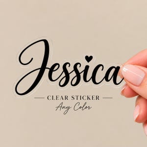 Puede incluir: Una pegatina transparente con el nombre Jessica escrito en una fuente cursiva negra. Un pequeño icono de corazón negro está colocado sobre la letra i. La pegatina tiene un borde troquelado que sigue la forma de las letras.