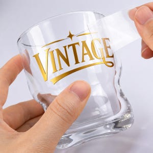 May include: A clear glass with a gold "VINTAGE" design. The glass is being held by a person, with a clear sticker being peeled off the glass. The design includes a star and a curved line.
