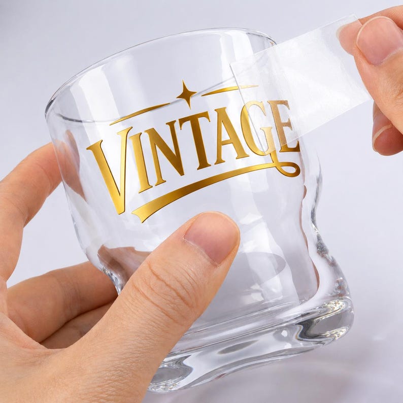 May include: A clear glass with a gold "VINTAGE" design. The glass is being held by a person, with a clear sticker being peeled off the glass. The design includes a star and a curved line.