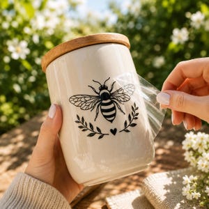 Puede incluir: Un recipiente cil&iacute;ndrico de cer&aacute;mica color crema con tapa de madera. Se est&aacute; aplicando un adhesivo de vinilo negro con forma de abeja rodeada por una rama con hojas y un peque&ntilde;o coraz&oacute;n, con una parte de la cinta de transferencia transparente retirada.