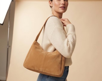 Könnte beinhalten: Eine beigefarbene Schultertasche aus Wildleder mit einem einzelnen Riemen, einer glatten Textur und einer vertikalen Naht in der Mitte. Die Tasche hat einen Reißverschluss oben und eine strukturierte, rechteckige Form mit abgerundeten Ecken.