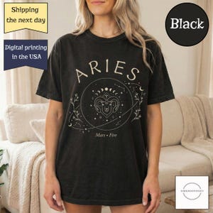 May include: Black t-shirt with the word "ARIES" in a cream color, along with a circular graphic featuring a ram's head, constellations, and the words "Mars • Fire". The shirt is a dark color.