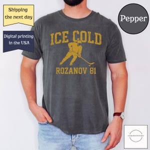 May include: Dark gray t-shirt with a distressed gold graphic that reads "ICE COLD" above a hockey player illustration and "ROZANOV 81". The shirt has a vintage look and feel. The shirt is a Pepper color.