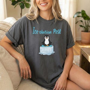 May include: Dark gray t-shirt with the text "Ice-olation Pose" in light blue. The shirt features a cartoon penguin sitting on an ice cube. The penguin is black and white with a yellow beak.