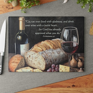 Könnte beinhalten: Dekoratives Schneidebrett mit einem Stillleben aus Wein, Brot, Käse und Trauben. Eine Weinflasche und ein Glas Rotwein sind zu sehen. Der Text "Go, eat your food with gladness, and drink your wine..." ist auf dem Brett aufgedruckt.