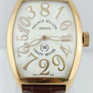 Puede incluir: Un reloj Franck Muller Crazy Hours dorado con una correa de cuero marrón. La esfera es blanca con grandes números dorados estilizados y las palabras "Franck Muller Geneve" y "Crazy Hours". El reloj tiene una forma rectangular curva única.
