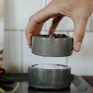 Könnte beinhalten: Eine Hand hält eine kleine, graue Keramikschale mit Pfefferkörnern über einer weiteren Schale mit Salz. Die Schalen stehen auf einem Metalltablett, im Vordergrund sind Chilischoten zu sehen. Die Szene deutet auf die Zubereitung von Speisen hin.