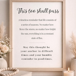 May include: A wooden framed print with the words "This too shall pass". The print is surrounded by decorative items, including a vase of flowers, books, and a lit candle, creating a calm and inspirational scene.