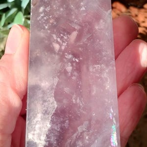 Könnte beinhalten: Ein hoher, rechteckiger Kristall mit polierter Oberfläche. Der Kristall hat eine blasslila Farbe mit weißen Einschlüssen und schillernden Reflexen. Der Kristall wird in einer Hand gehalten.
