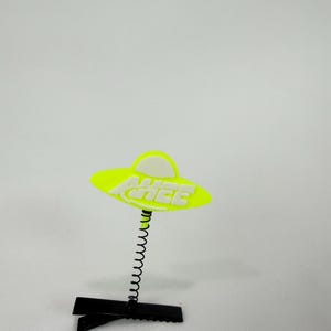 May include: A neon yellow UFO-shaped hair clip with the word "AHHEE" in white letters. The clip is attached to a black metal clip with a black spring. The background is plain white.
