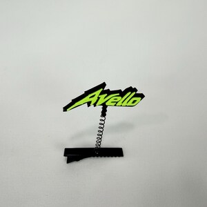 May include: A black and neon green Avello logo on a spring clip. The logo is in a stylized font, with the word "Avello" in black and neon green. The clip is black and has a spring coil.