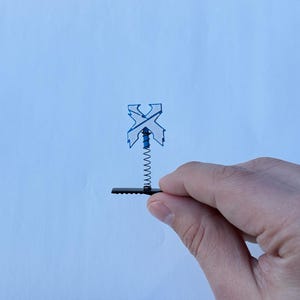 May include: A blue and black mechanical component held by a hand. The component features a spring and a stylized "X" design. The background is a light blue surface.