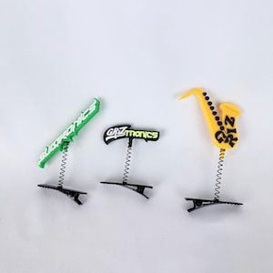 May include: Three novelty clips with spring attachments. One clip has a green sign with the text "GRZTRONICS", another has a black sign with the text "GRZTRONICS", and the third is a yellow saxophone with the text "212".