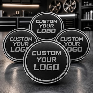 May include: A set of four circular black wheel centre caps with a silver metallic border. Each cap features the embossed silver text CUSTOM YOUR LOGO centred on the face. The caps are arranged in a cluster, displaying a matte finish and clean lines.