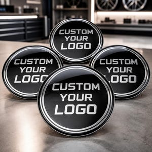 May include: Four circular black emblems with a metallic silver border and the text CUSTOM YOUR LOGO embossed in silver lettering. The emblems feature a glossy finish and are arranged in a cluster, showcasing a reflective surface and clean design.