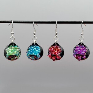 May include: Four pairs of dangle earrings with silver hooks. Each earring features a black, rounded base filled with colorful beads. The beads are in shades of green, blue, red, and purple.
