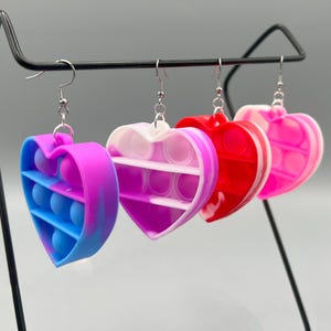 May include: Four heart-shaped earrings in a row, each with a different color scheme. The first is blue and purple, the second is white and purple, the third is red, and the fourth is pink. Each earring has a silver hook.