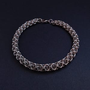 Könnte beinhalten: Ein silberfarbenes Kettenarmband mit einem gewebten Design. Das Armband ist kreisförmig und liegt auf einem dunklen Hintergrund. Der Verschluss ist oben sichtbar. Länge ca. 20 cm.