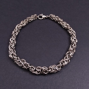 May include: A silver-colored Byzantine chain bracelet. The bracelet is made of interlocking metal links, creating a textured, woven appearance. The clasp is a small, secure lobster claw style. The bracelet is displayed on a dark background.