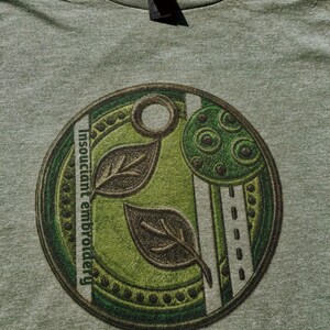 May include: A heathered olive green t-shirt featuring a circular graphic print. The design includes two dark leaves, a textured green circle, and a vertical white stripe with dashed lines. The text "Insouciant embroidery" is printed vertically.