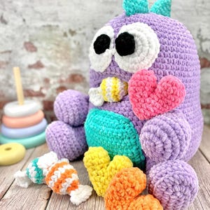 Puede incluir: Un gran monstruo morado hecho a mano con ganchillo, con ojos blancos, pupilas negras y detalles turquesa. Tiene una barriga turquesa y está rodeado de corazones y caramelos de ganchillo de varios colores. Una pila de anillos de colores está en el fondo.