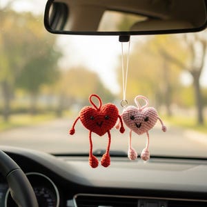 May include: Two crocheted heart-shaped car charms. One is red, the other is pink. Each has arms, legs, and a smiling face. They hang from the rearview mirror in a car, adding a touch of handmade charm to the vehicle's interior.