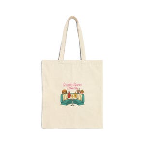 May include: Cream-colored tote bag featuring a retro diner scene with the text "Greasy Spoon Dreams." The illustration includes a burger, fries, and a milkshake. The bag has a simple design with two handles.