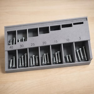 May include: A gray organizer with labeled compartments holding various sizes of silver bolts. The compartments are labeled with numbers from 5 to 40. The organizer is made of a solid, matte material.