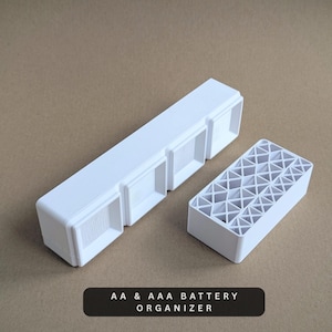 May include: A white AA and AAA battery organizer. The organizer has two parts: a long rectangular holder with three compartments and a smaller rectangular holder with a grid pattern. The text "AA & AAA BATTERY ORGANIZER" is at the bottom.