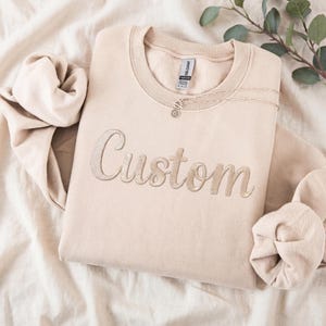 May include: A beige crew neck sweatshirt embroidered with the word "Custom" in a lighter beige thread. A delicate necklace with a circular pendant rests on the sweatshirt. The sweatshirt is folded on a soft, textured surface.