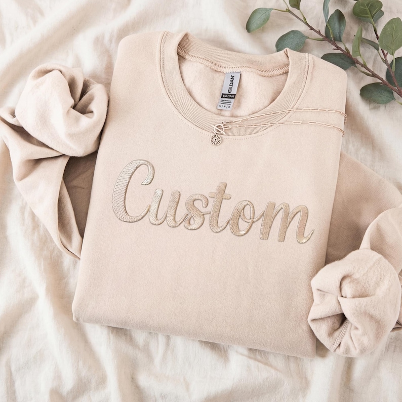 May include: A beige crew neck sweatshirt embroidered with the word "Custom" in a lighter beige thread. A delicate necklace with a circular pendant rests on the sweatshirt. The sweatshirt is folded on a soft, textured surface.