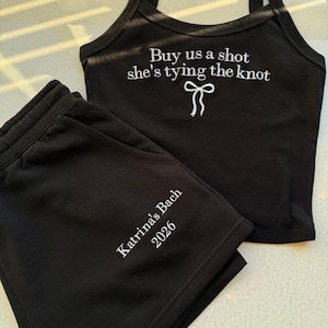 May include: Black matching tank top and shorts set. The tank top has the text "Buy us a shot she's tying the knot" with a bow graphic. The shorts are embroidered with "Katrina's Bach 2026".