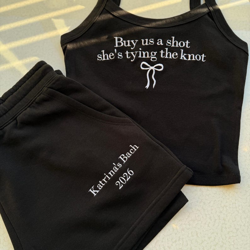 May include: Black matching tank top and shorts set. The tank top has the text "Buy us a shot she's tying the knot" with a bow graphic. The shorts are embroidered with "Katrina's Bach 2026".