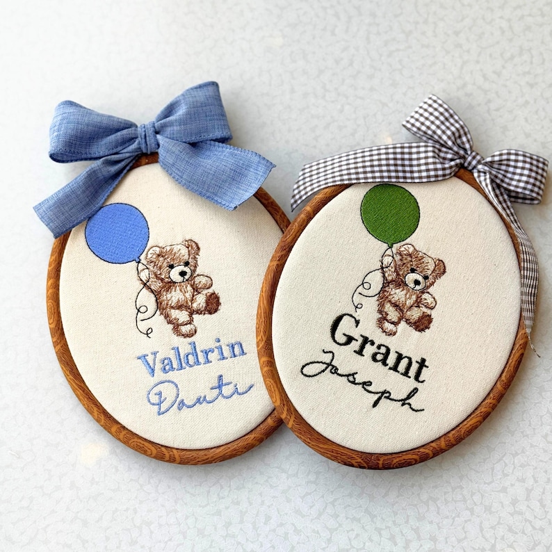 May include: Two embroidered oval hoops with teddy bears holding balloons. One has a blue balloon and bow, the other a green balloon and gingham bow. Names are embroidered below the bears. The hoops have wooden frames.