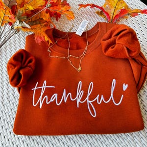 May include: An orange sweatshirt embroidered with the word "thankful" in white script. A gold necklace with a heart pendant and two orange scrunchies are also visible. Autumn leaves are arranged around the sweatshirt.